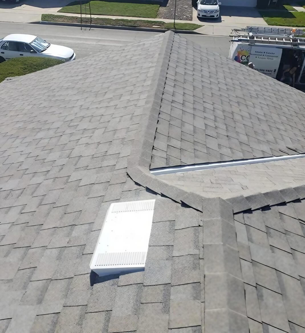 Aerial view of a gray shingled roof with a white vent on a sunny day, showcasing a well-maintained, pitched design.