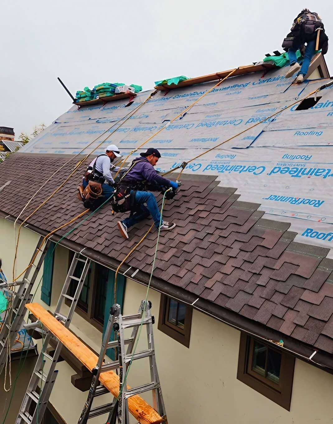 Three workers are installing roofing shingles on a house, using safety ropes, ladders, and scaffolding amid overcast skies.